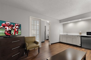 A modern living room with a flat-screen TV, a dark wood cabinet, a beige armchair, and a kitchen area with white cabinets and a stainless steel refrigerator. - Red Roof Inn & Suites Savannah Airport (Pooler)