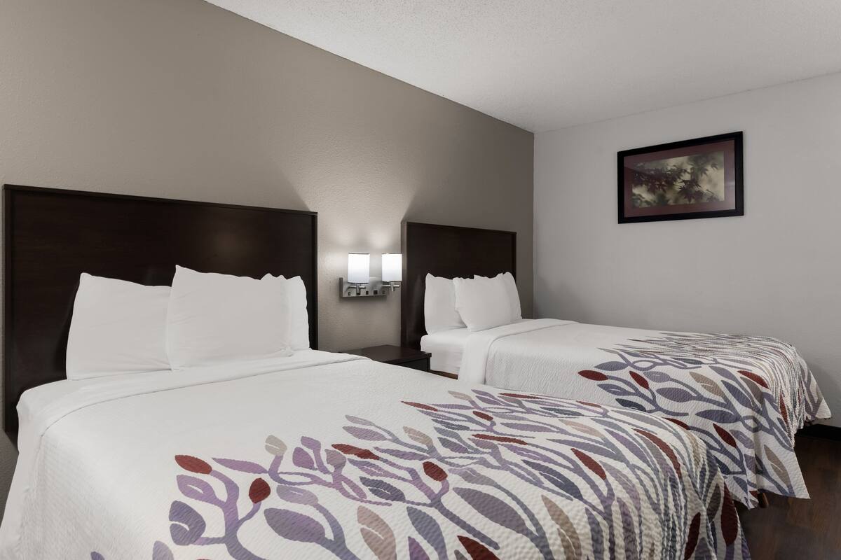 A hotel room with two beds, each with a dark headboard and white bedding. There is a wall-mounted light fixture above the beds and a framed picture on the wall.