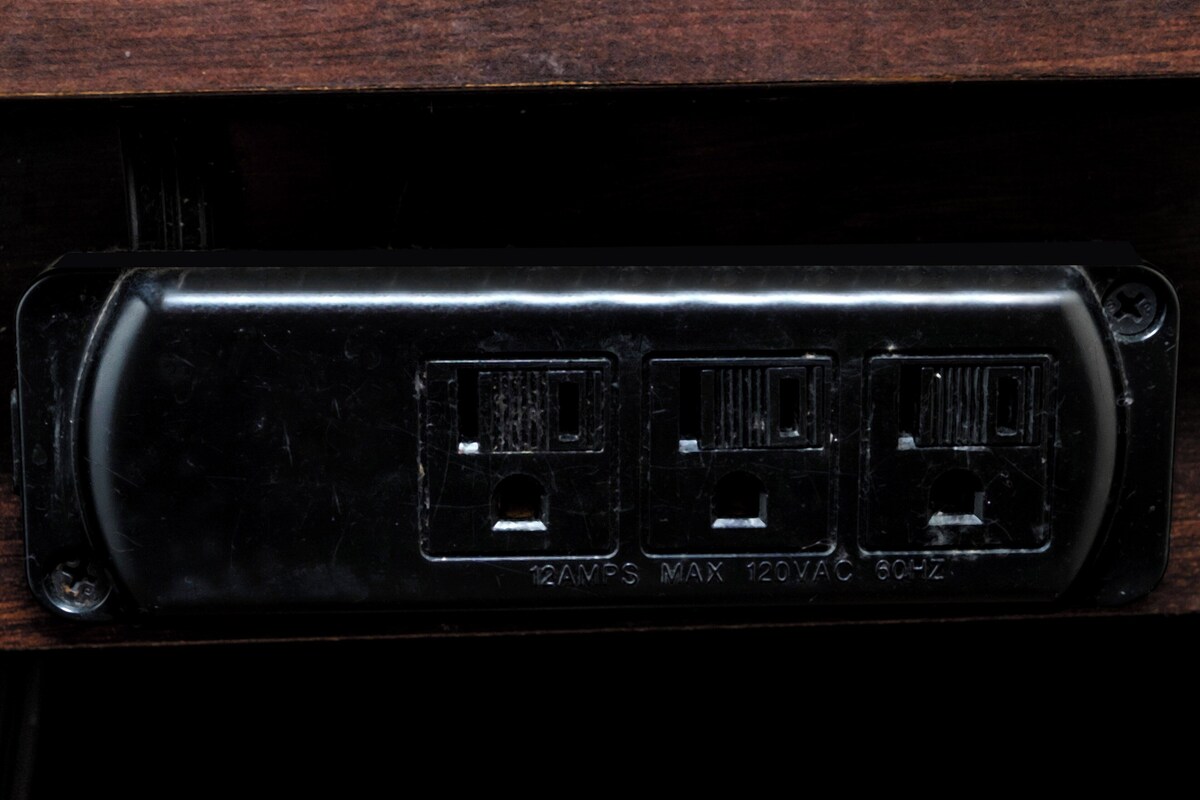 Un enchufe eléctrico negro con tres tomas, etiquetado como '12AMP MAX 120VAC 60HZ'.