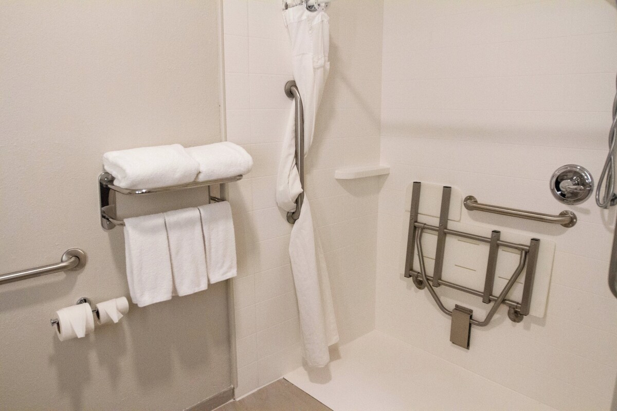 A bathroom with a toilet paper holder, a towel rack with towels, a shower area with a grab bar, and a hand dryer.