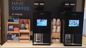 A coffee station with three espresso machines, a display of coffee pods, and a sign that reads "MUST. HAVE. COFFEE." - Holiday Inn Express Oneill by IHG (O'Neill)