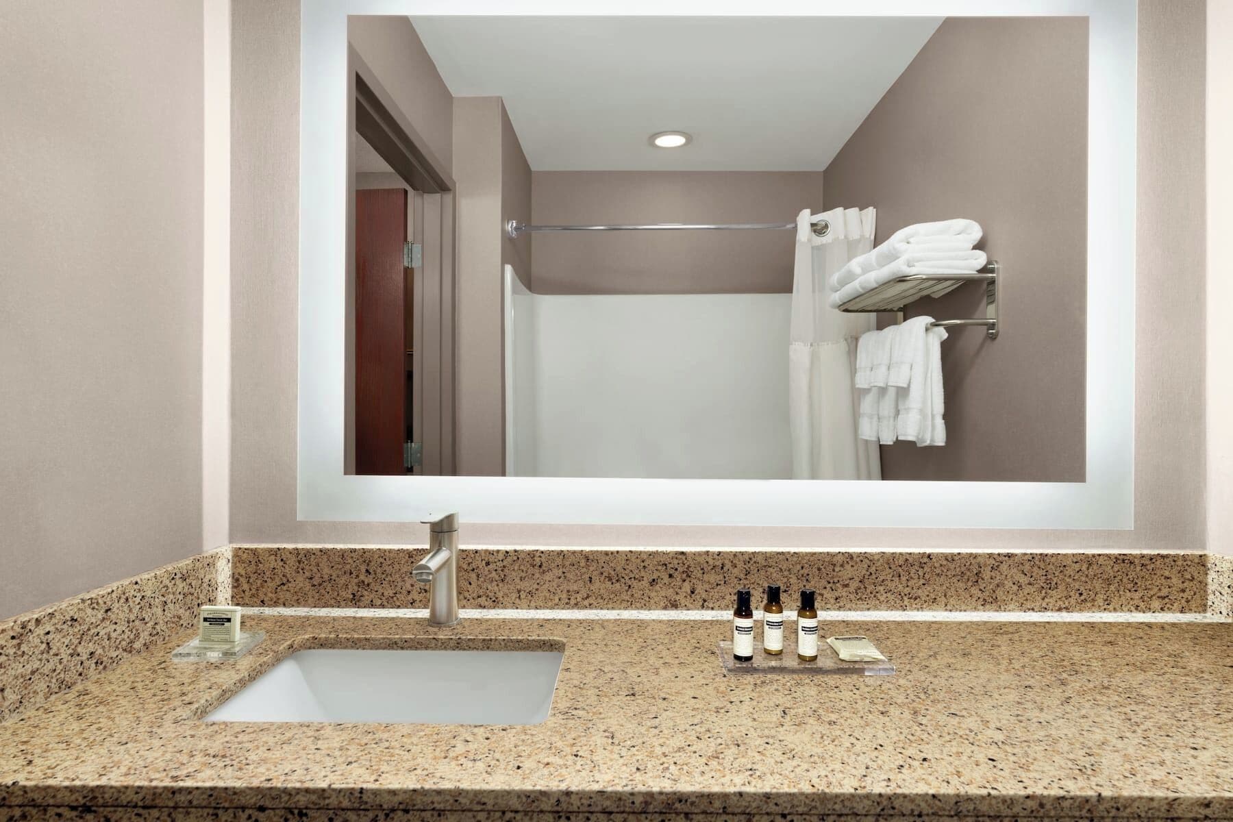 Un baño con un espejo grande, un lavabo con encimera de granito y un perchero con toallas dobladas.