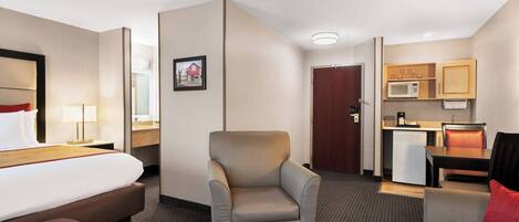 A hotel room with a bed, a chair, a desk, a microwave, and a small kitchenette.