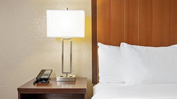 Quarto de hotel com cabeceira de madeira, mesa de cabeceira com abajur, telefone e cama bem arrumada.