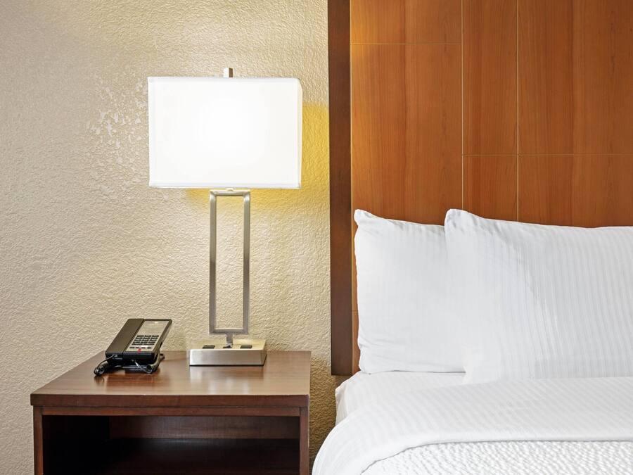 Quarto de hotel com cabeceira de madeira, mesa de cabeceira com abajur, telefone e cama bem arrumada.