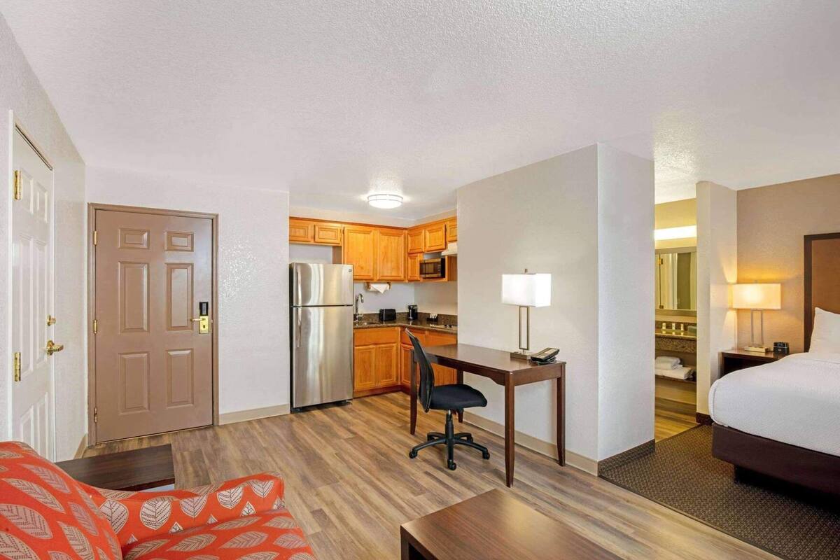 A hotel room with a bed, desk, chair, kitchenette, and a door.