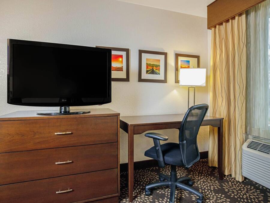 Habitación de hotel con televisión sobre un mueble de madera, escritorio con silla, lámpara y cuadros en la pared.