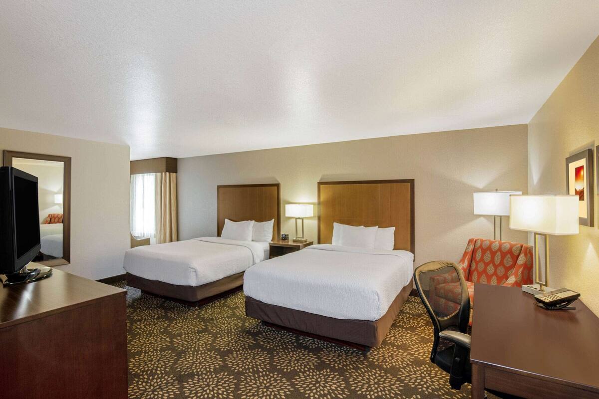 A hotel room with two beds, a desk, a chair, a TV, and a lamp.