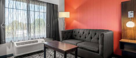Une chambre d’hôtel avec un canapé gris, une table en bois, un lampadaire et une grande fenêtre avec des rideaux.