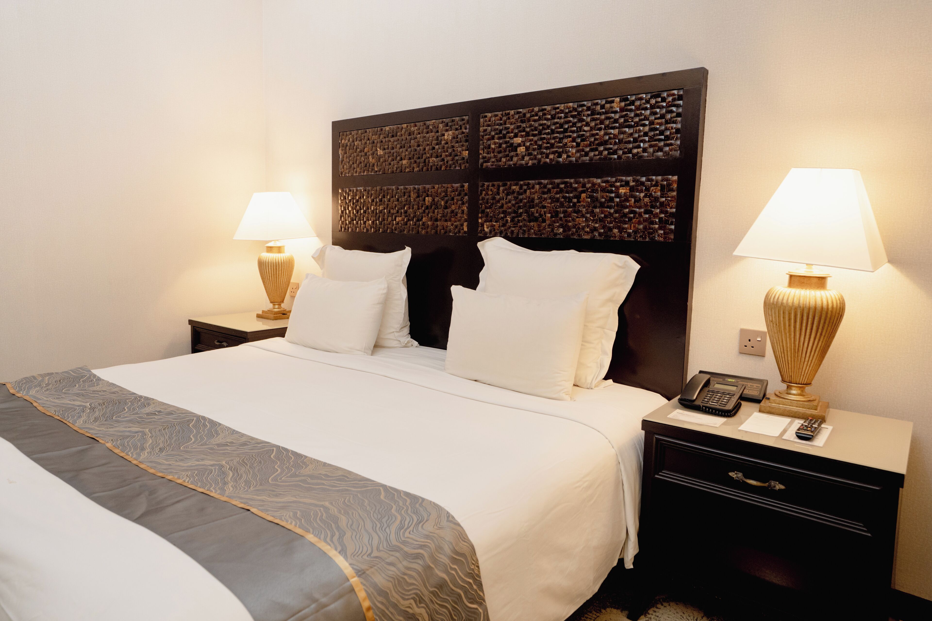 Una cama bien hecha con sábanas blancas, una manta estampada y un cabecero oscuro con un diseño de mosaico. Se observan dos mesitas de noche, una con lámparas y un teléfono.