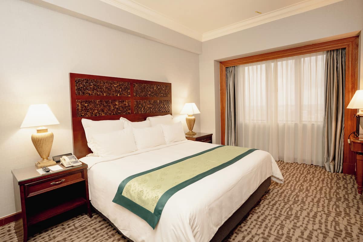 A hotel room with a large bed, two bedside tables with lamps, a decorative headboard, and a window with curtains.