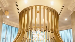Un espace salon moderne aux lignes courbes, avec une table centrale, des sièges recouverts d’un tissu clair et un élément décoratif au plafond.