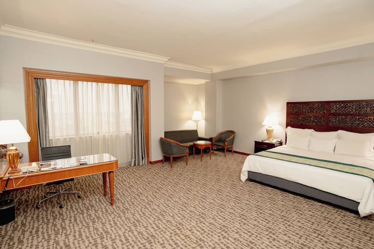 A hotel room with a large bed, a desk with a computer, two armchairs, a small table, and a window with curtains.