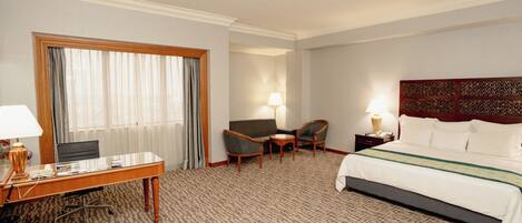 Habitación de hotel con una cama grande, un escritorio con computadora, dos sillones, una mesita y una ventana con cortinas.