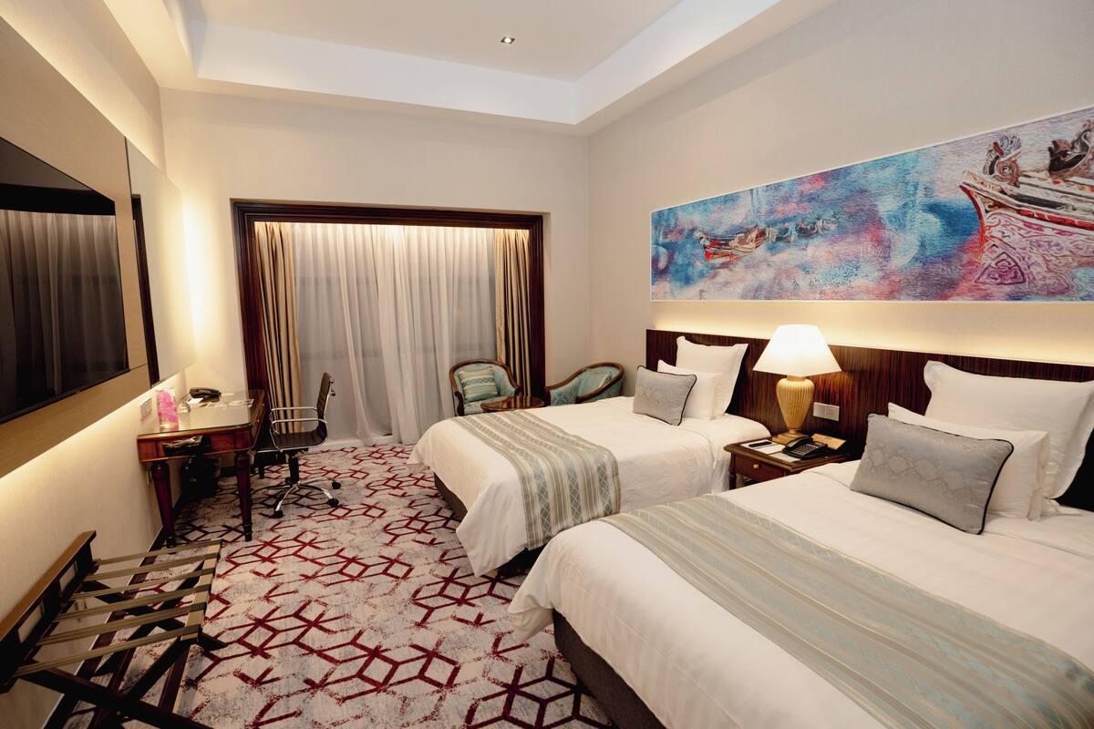 A hotel room with two beds, a desk, a chair, a large painting on the wall, and a patterned carpet.