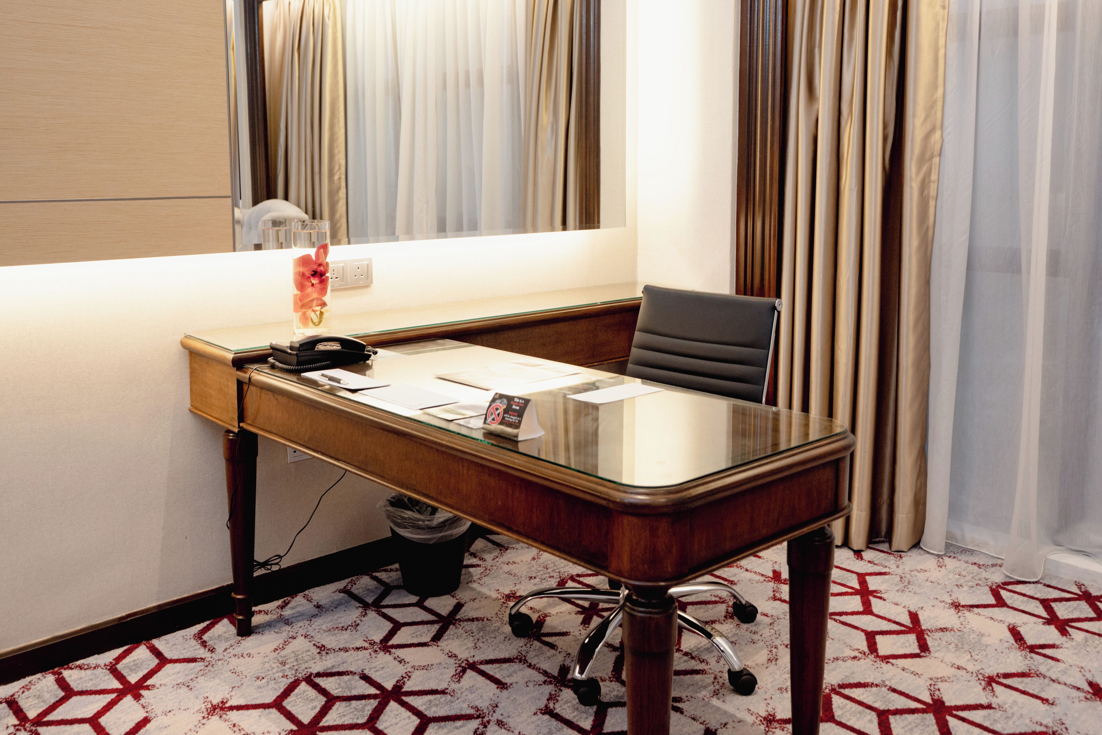 A hotel room with a wooden desk, a chair, a glass-top table, a telephone, and a small trash bin.