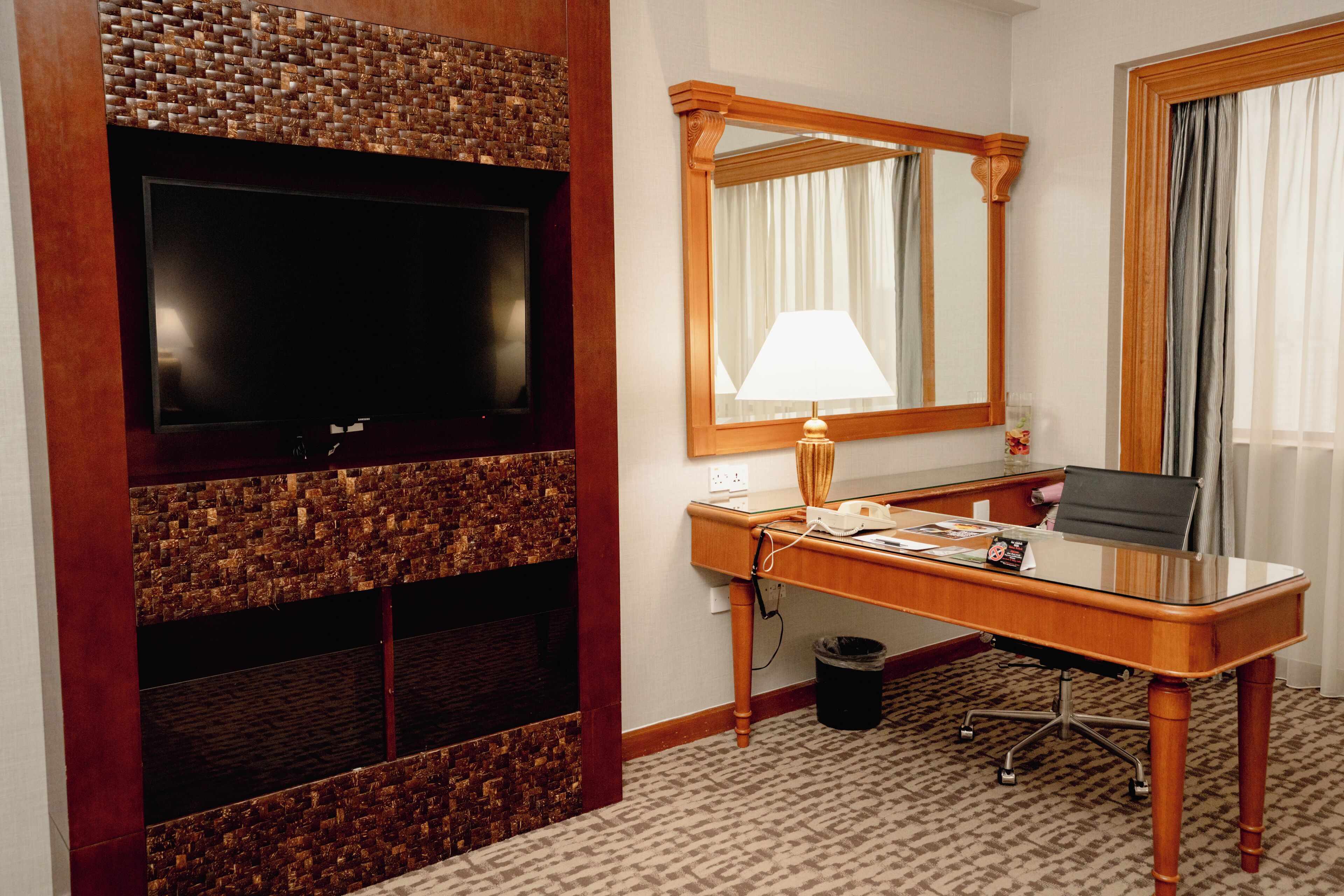 A hotel room with a flat-screen TV mounted on a wooden wall, a desk with a lamp, a chair, and a large mirror.