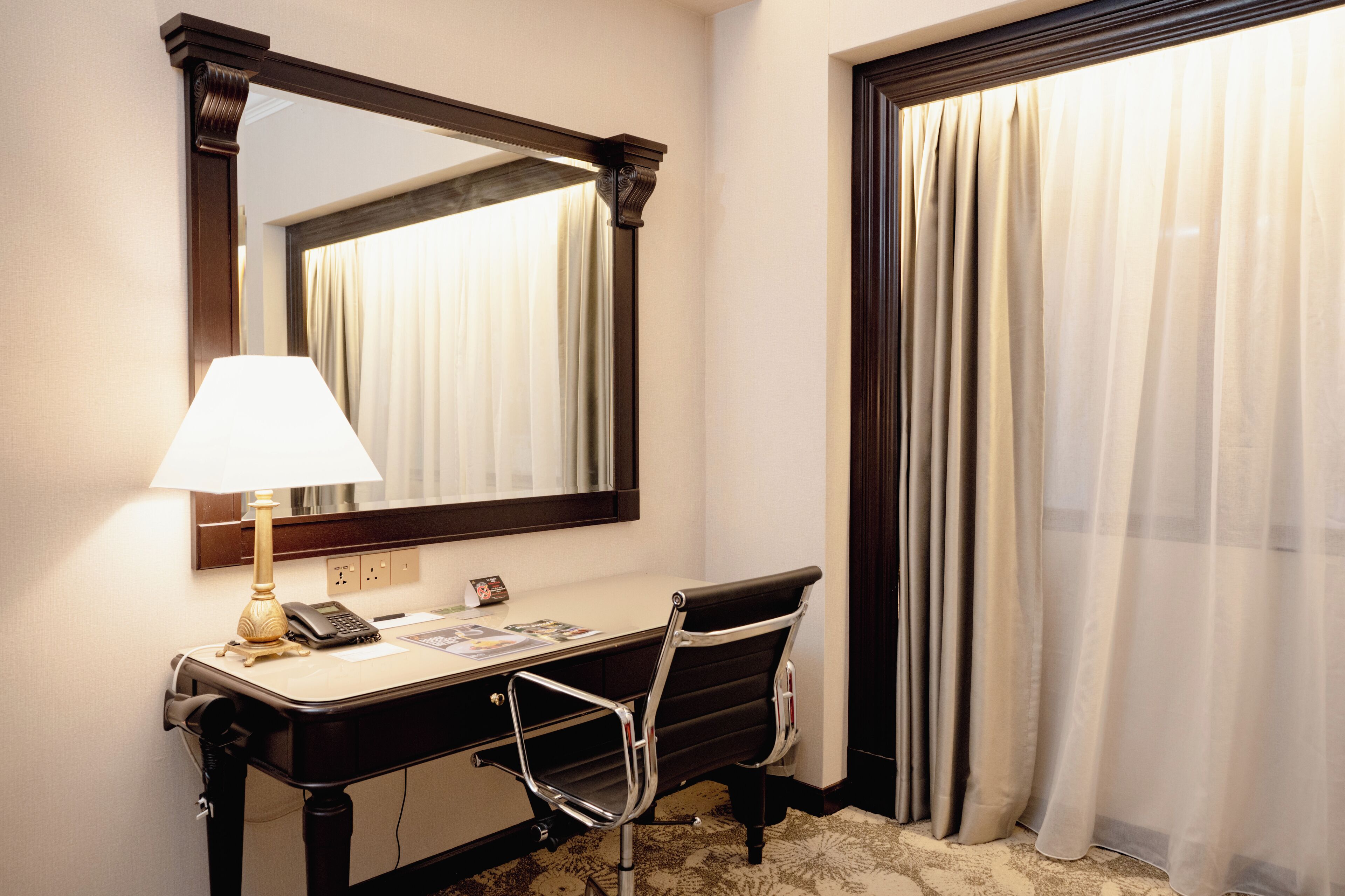 A hotel room with a desk, chair, lamp, telephone, and a large mirror.