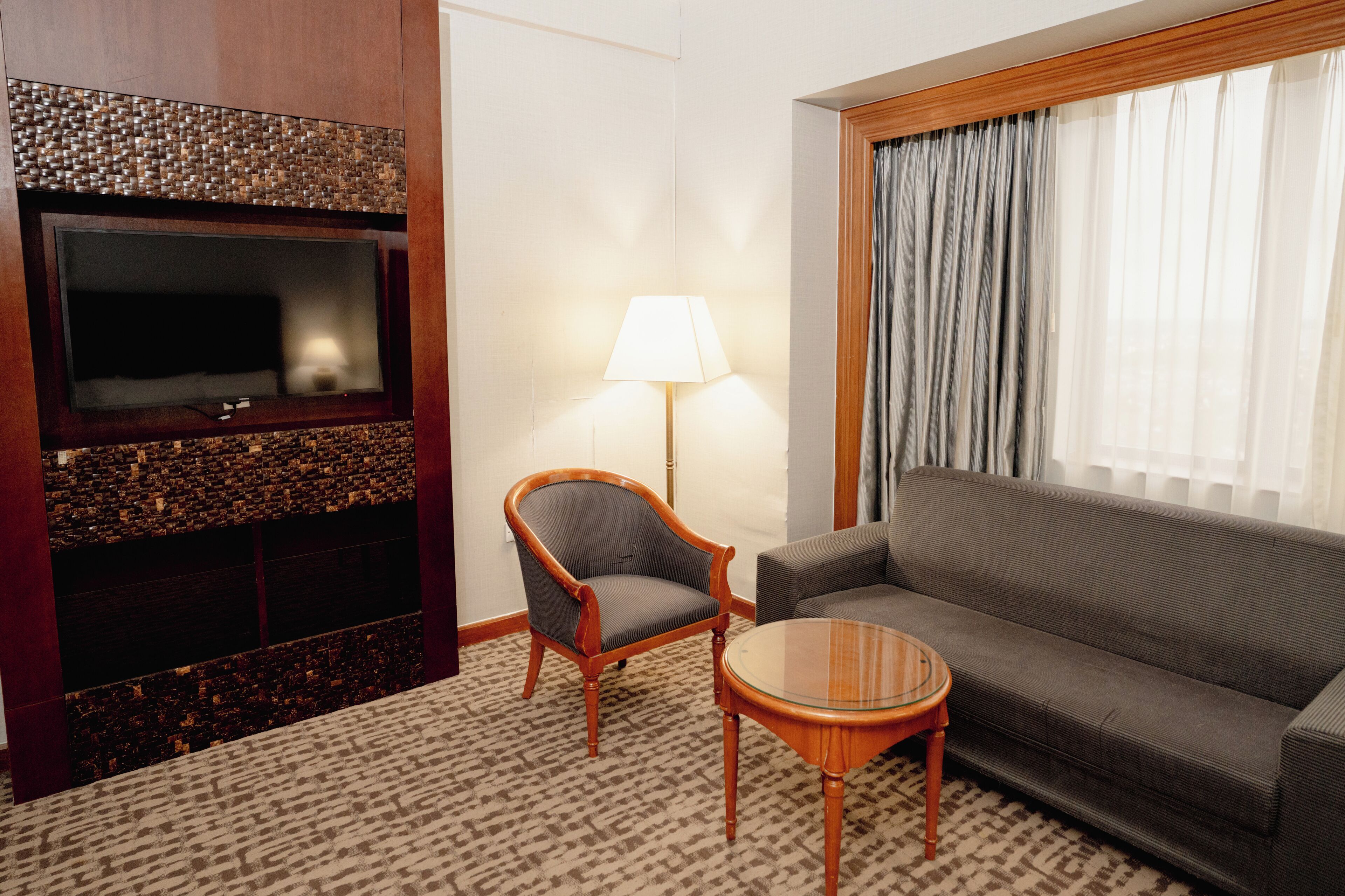 A hotel room with a flat-screen TV, a wooden chair, a sofa, a lamp, and a patterned carpet.