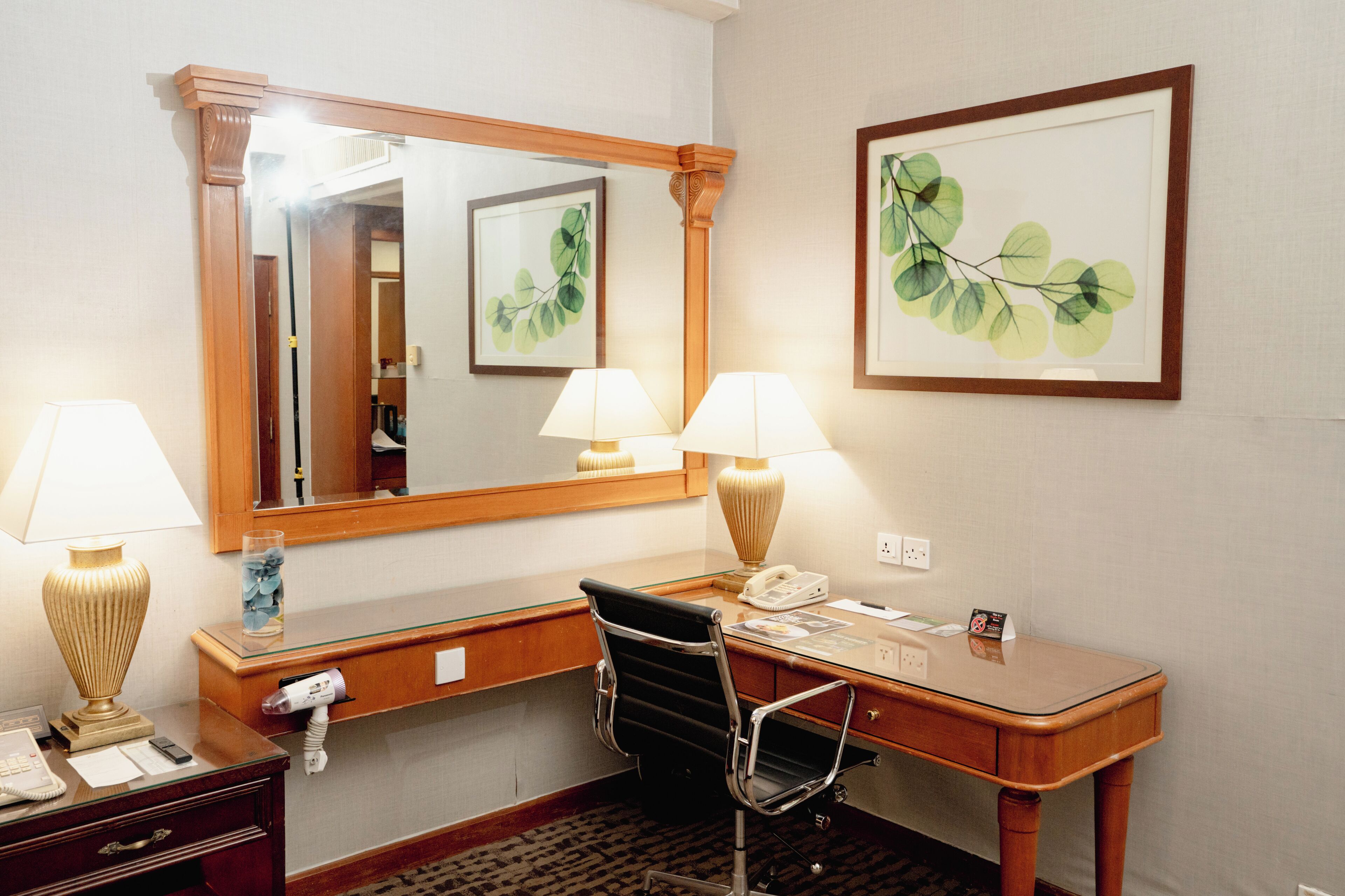 Habitación de hotel con un escritorio de madera, dos lámparas, una silla, un espejo y cuadros en la pared.