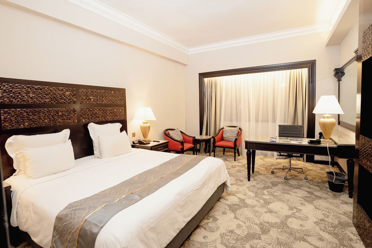 A hotel room with a large bed, a desk with a computer, two chairs, a window with curtains, and a lamp on each side of the bed.