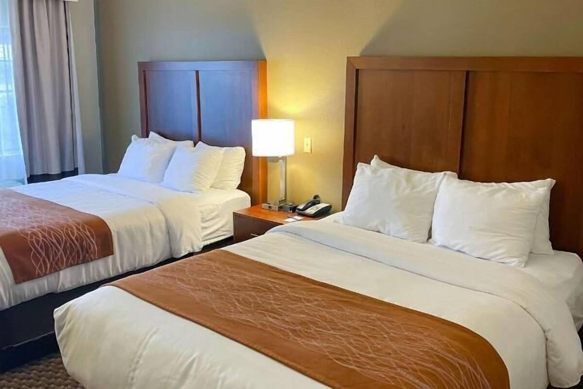 A hotel room with two beds, a nightstand, a lamp, and a window with curtains.