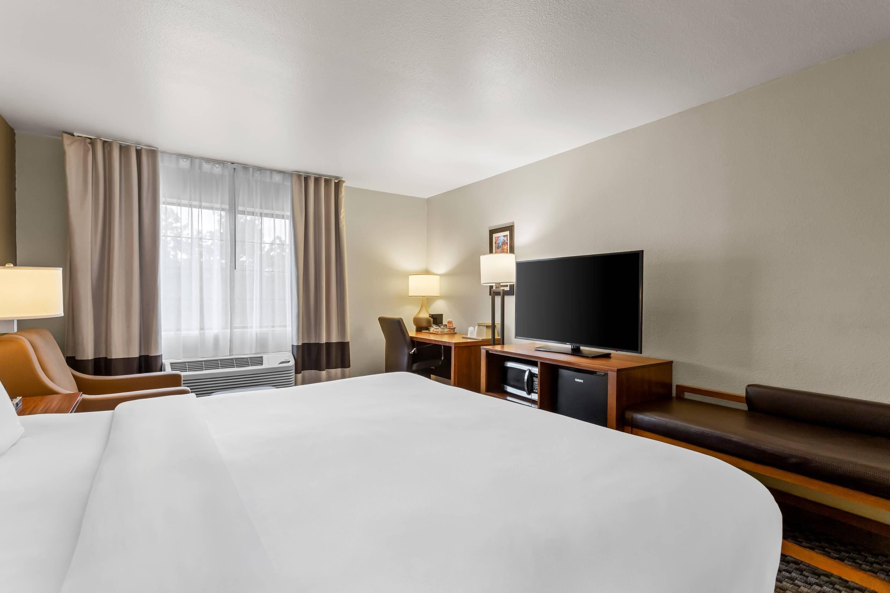 A hotel room with a large bed, a desk with a flat-screen TV, a chair, a sofa, and a window with curtains.