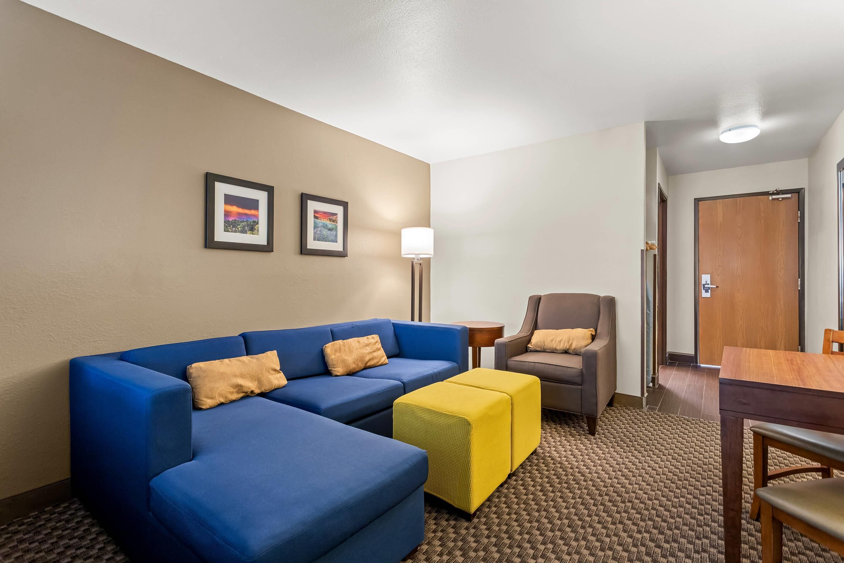A hotel room with a blue sofa, a yellow ottoman, a brown armchair, a small table, and two framed pictures on the wall.