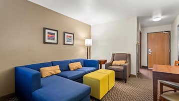 A hotel room with a blue sofa, a yellow ottoman, a brown armchair, a small table, and two framed pictures on the wall.