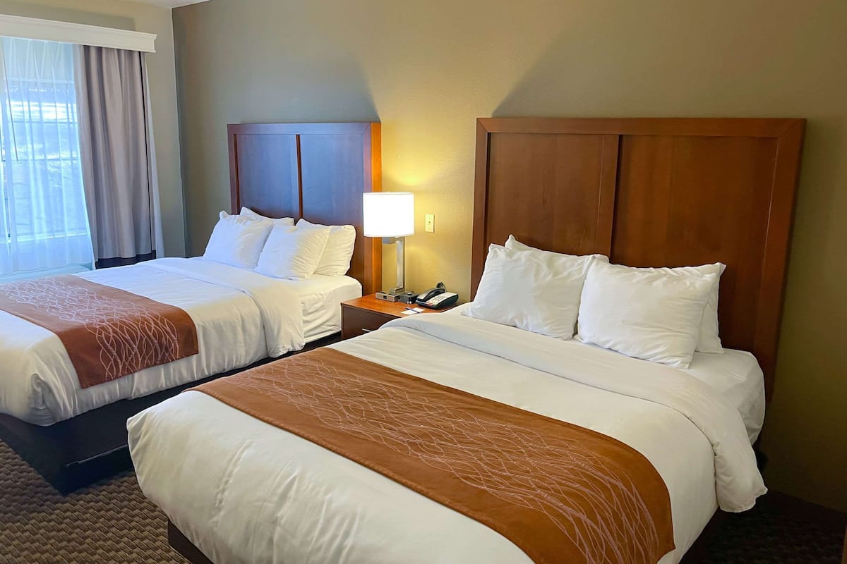 A hotel room with two beds, a nightstand with a lamp, and a view of the outdoors through a window.
