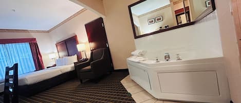A hotel room with a bed, a chair, a desk, a desk lamp, a mirror, and a bathroom with a bathtub and sink.