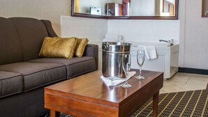 A hotel room with a brown sofa, a wooden coffee table with a metal bucket and two wine glasses, a large mirror, and a white bathroom with a sink and towel. - Comfort Suites Portland Southwest (Portland)