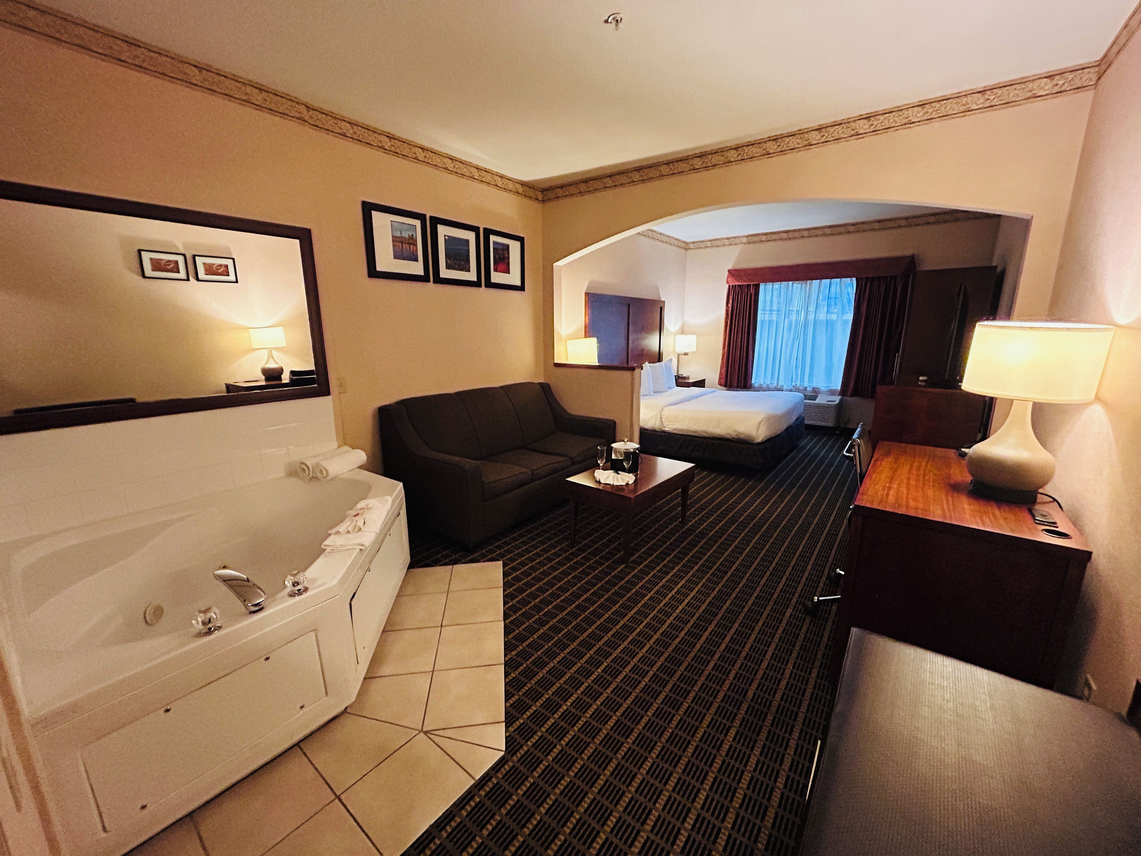 A hotel room with a large bed, a sofa, a desk, a lamp, a bathroom with a bathtub, and a shower.