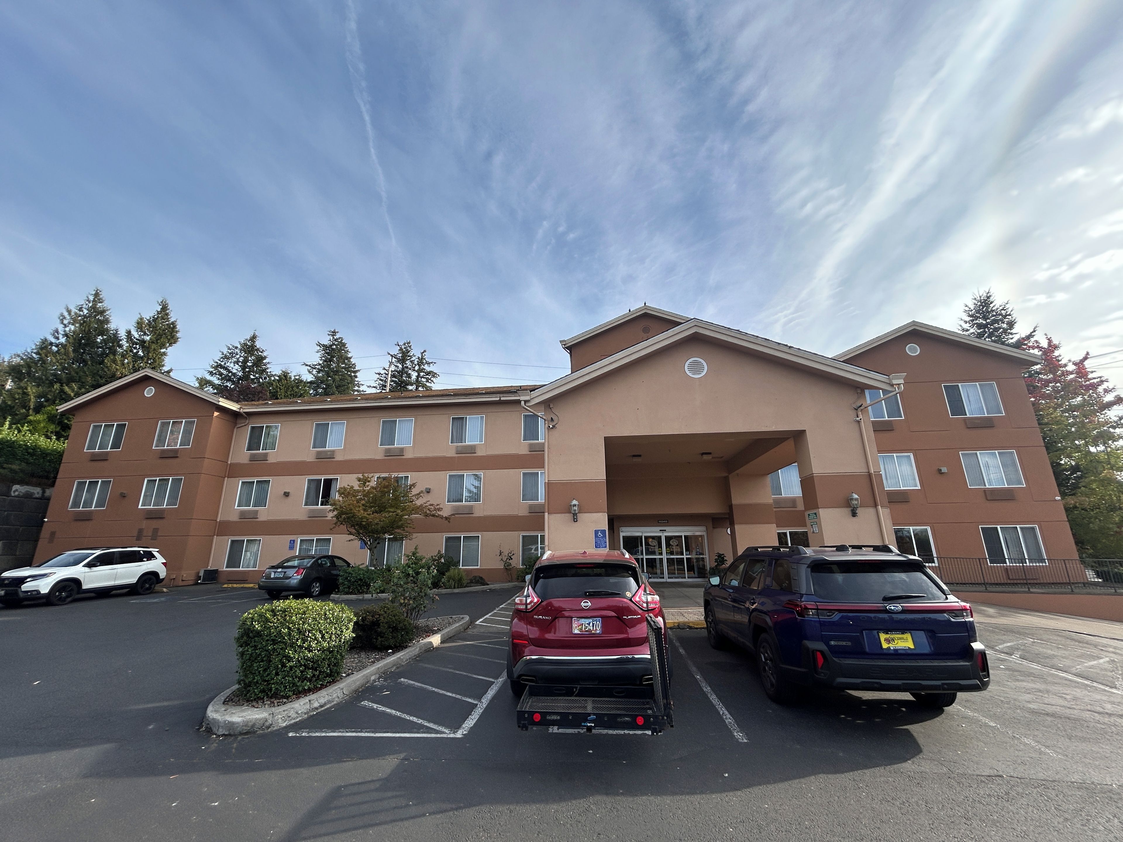 A multi-story hotel with a central entrance, parking lot, and surrounding trees.