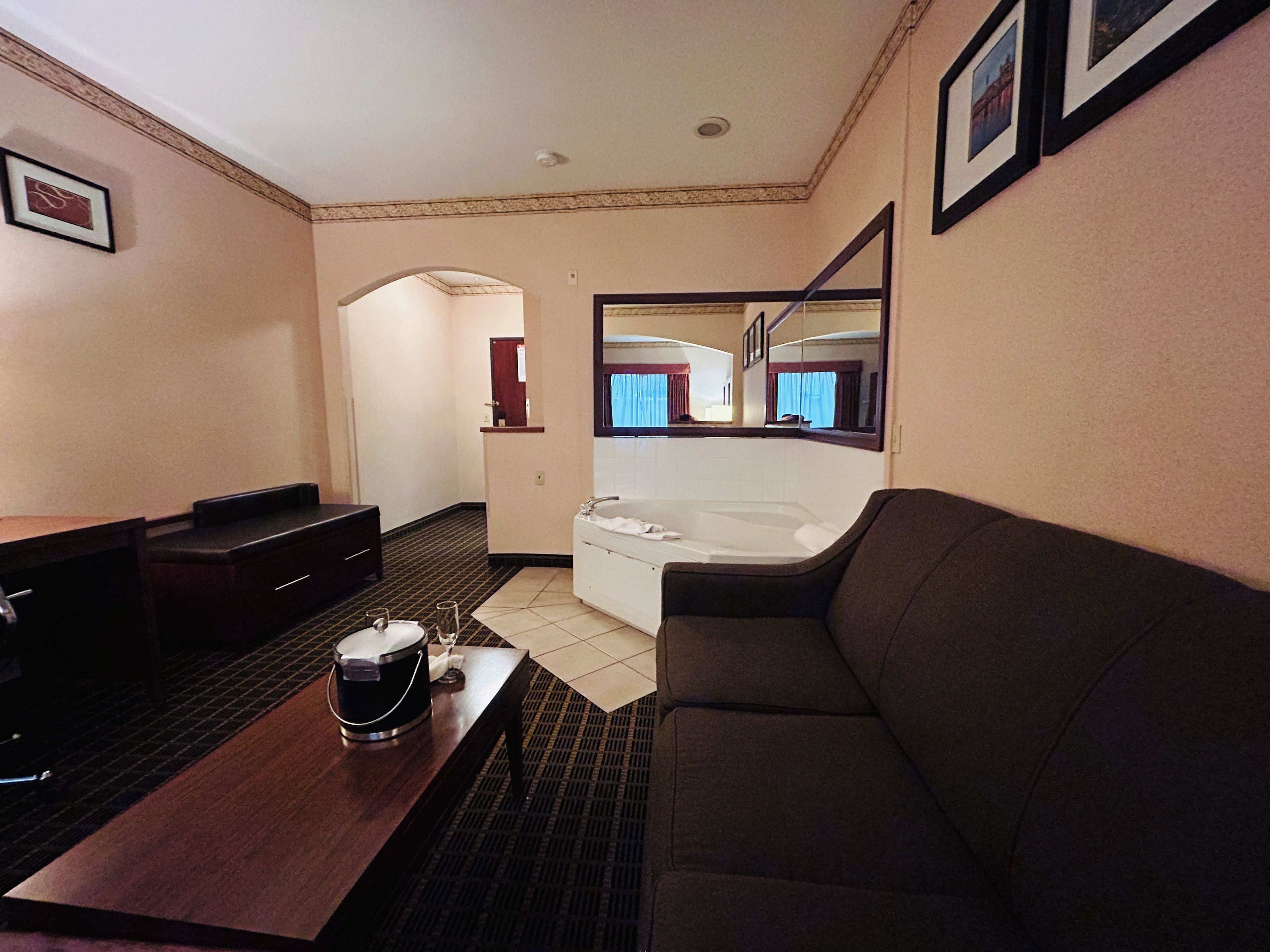 Habitación de hotel con sofá, una mesita con un vaso y un cubo, bañera y un espejo que refleja una ventana con cortinas.