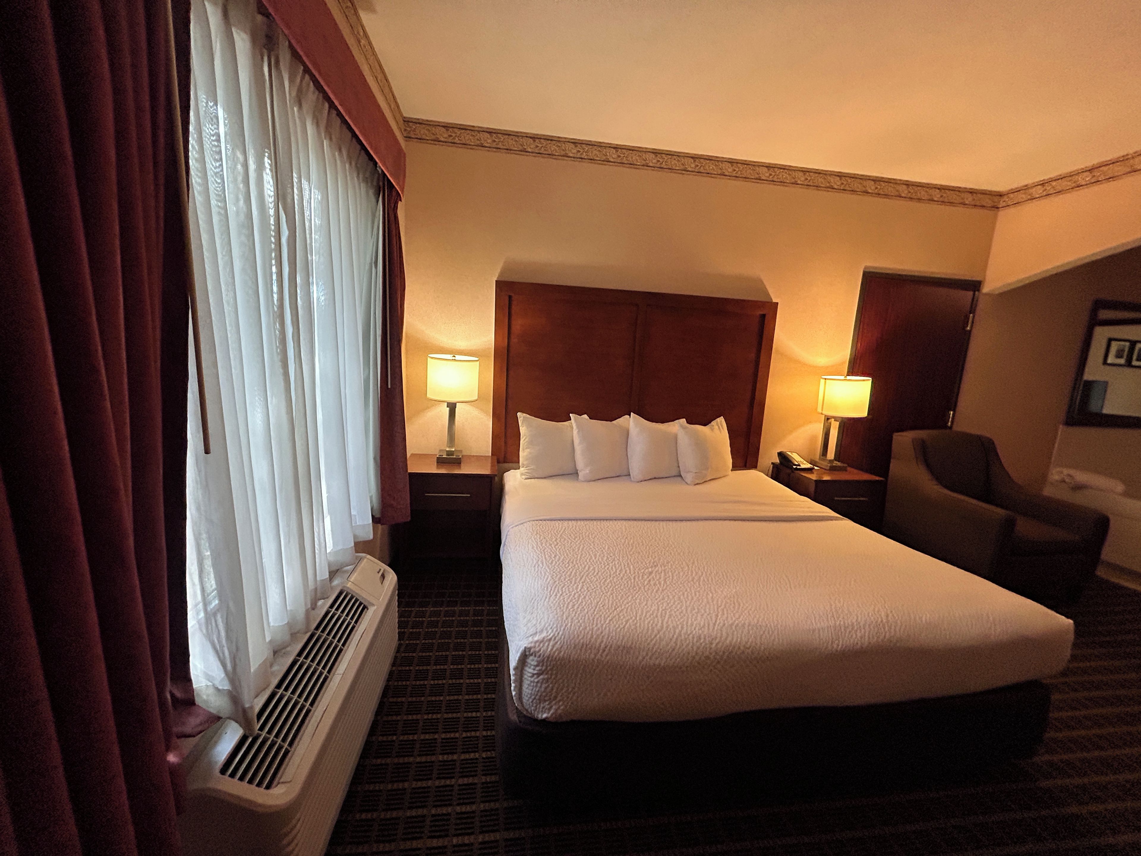 A hotel room with a large bed, two bedside lamps, a chair, and a window with curtains.
