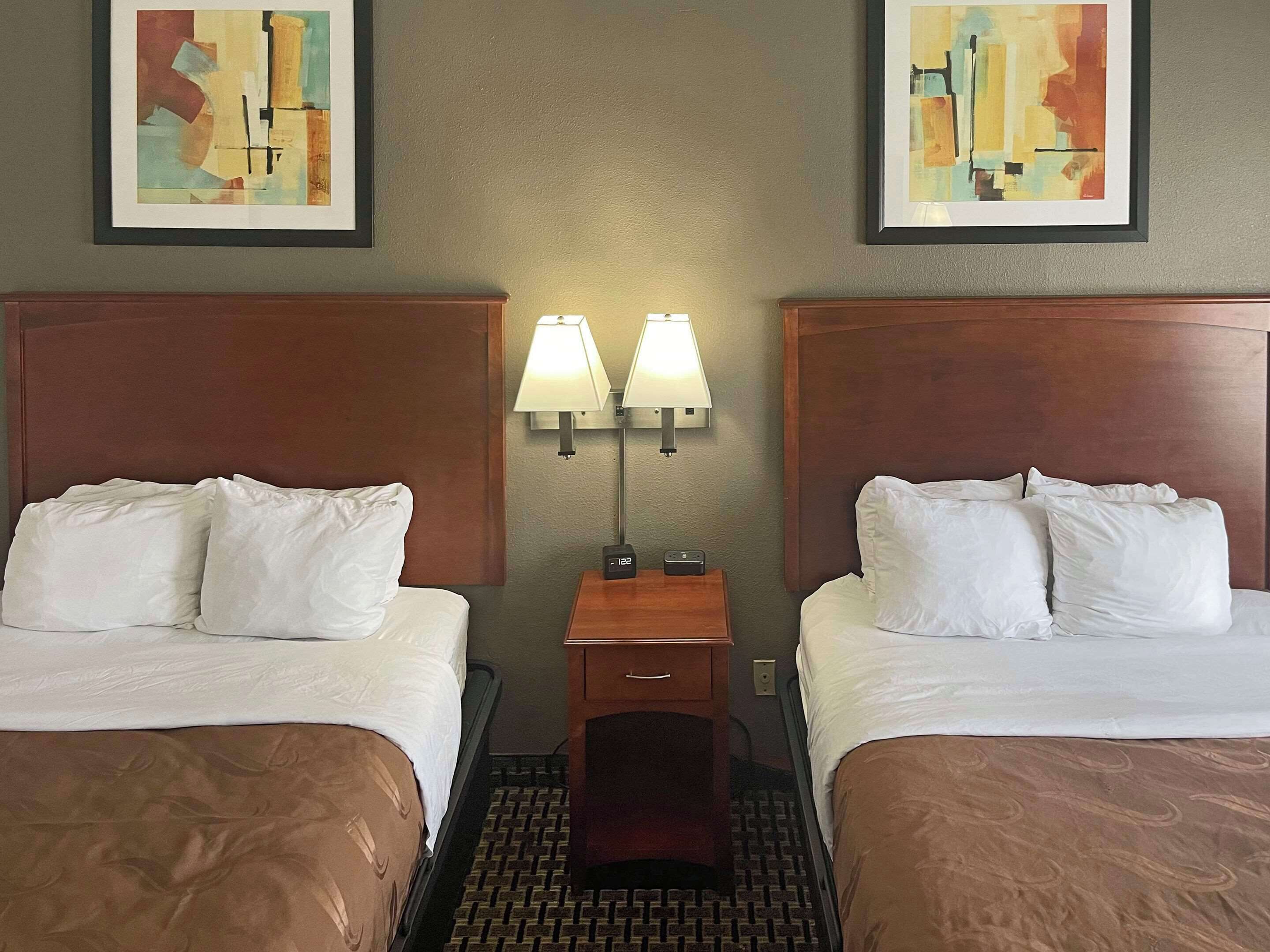 Una habitación de hotel con dos camas, una mesita de noche con lámparas y cuadros abstractos en las paredes.