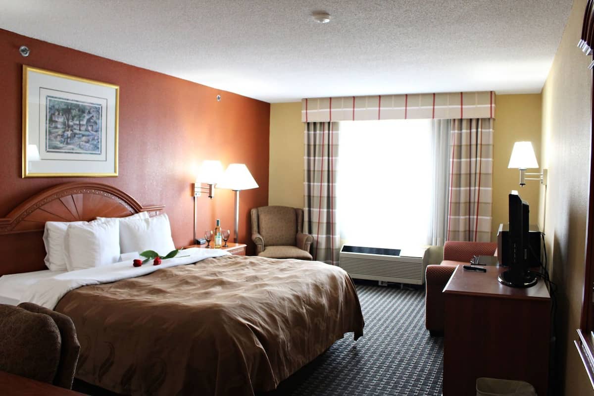 A hotel room with a bed, two bedside lamps, a chair, a desk with a computer, and a window with curtains.