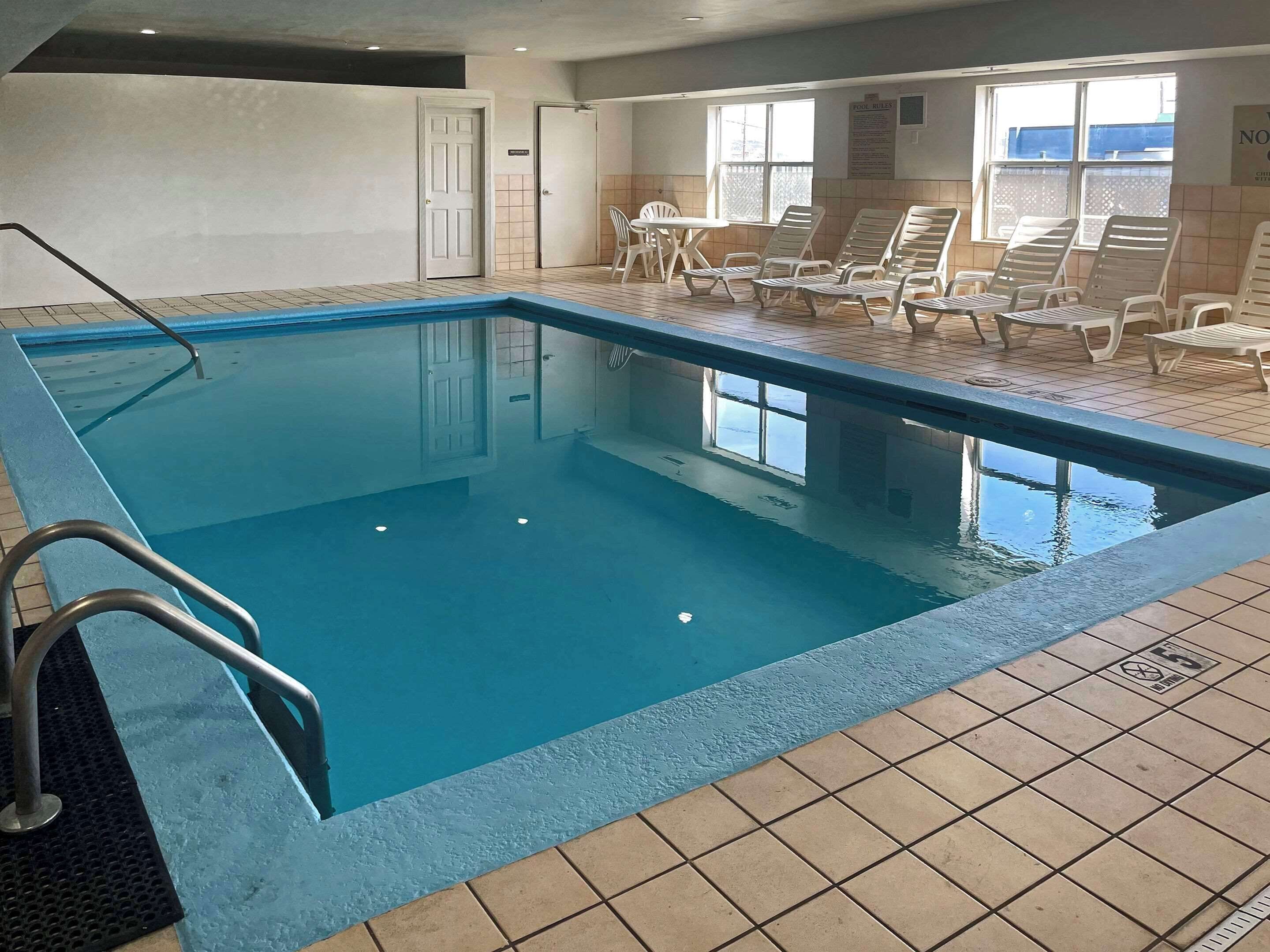 An indoor swimming pool with tiled flooring, a metal pool ladder, and white lounge chairs.