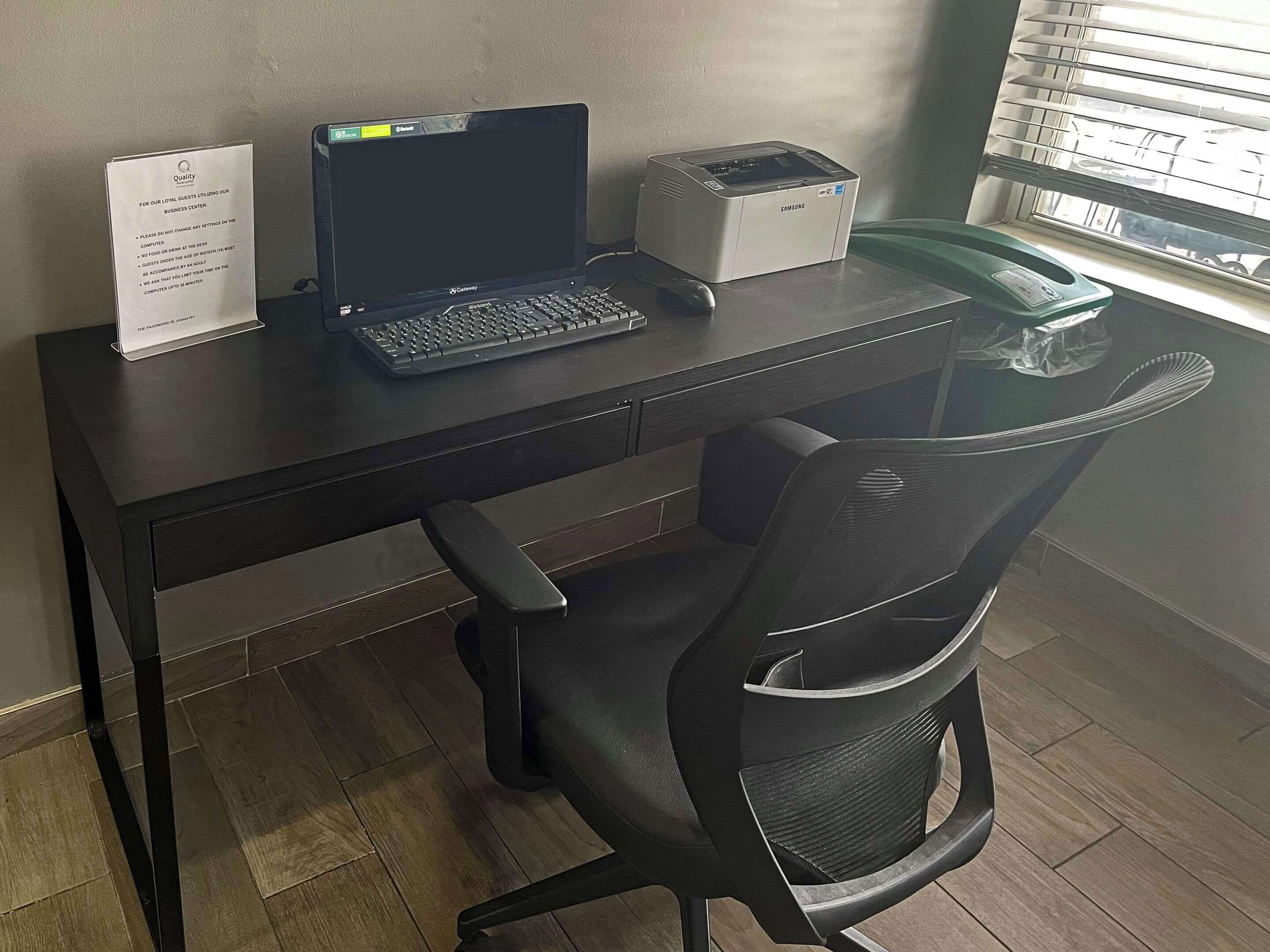 A desk with a laptop, a Samsung printer, and a telephone. There is a sign on the desk and a chair in front of it.