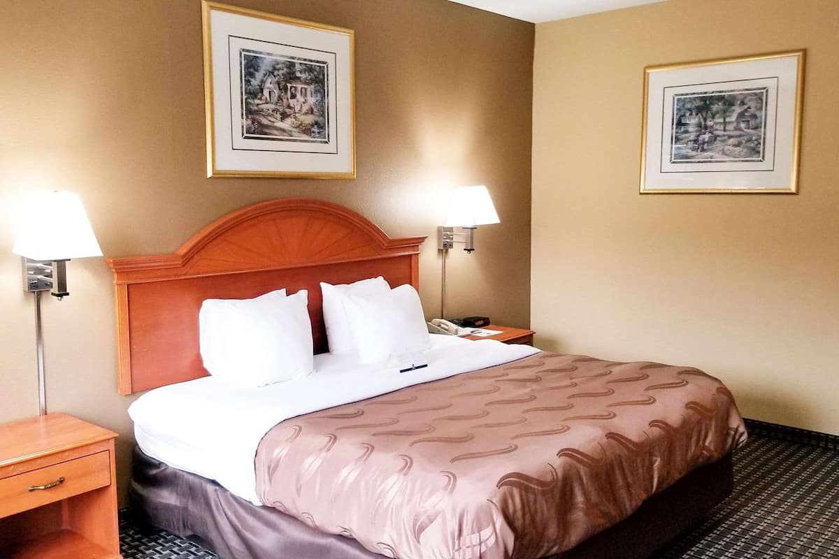 A hotel room with a wooden headboard, a bed with white linens, two bedside lamps, a nightstand, and two framed pictures on the wall.
