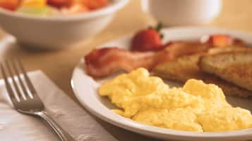 A breakfast setting with scrambled eggs, bacon, toast, a fruit bowl, and a coffee cup on a table.