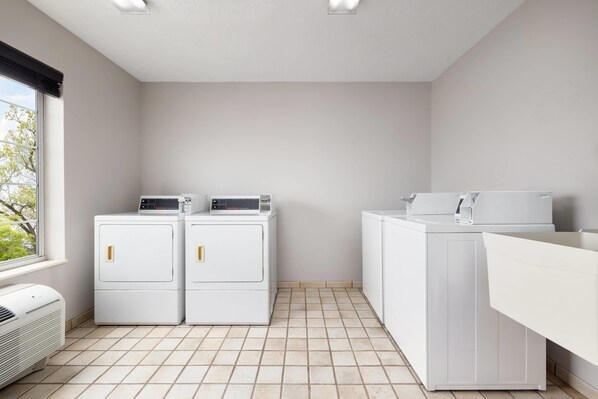 A laundry room with two front-loading washers and dryers, a white sink, and a window with blinds. - Country Inn & Suites by Radisson, Springfield, OH (Springfield)