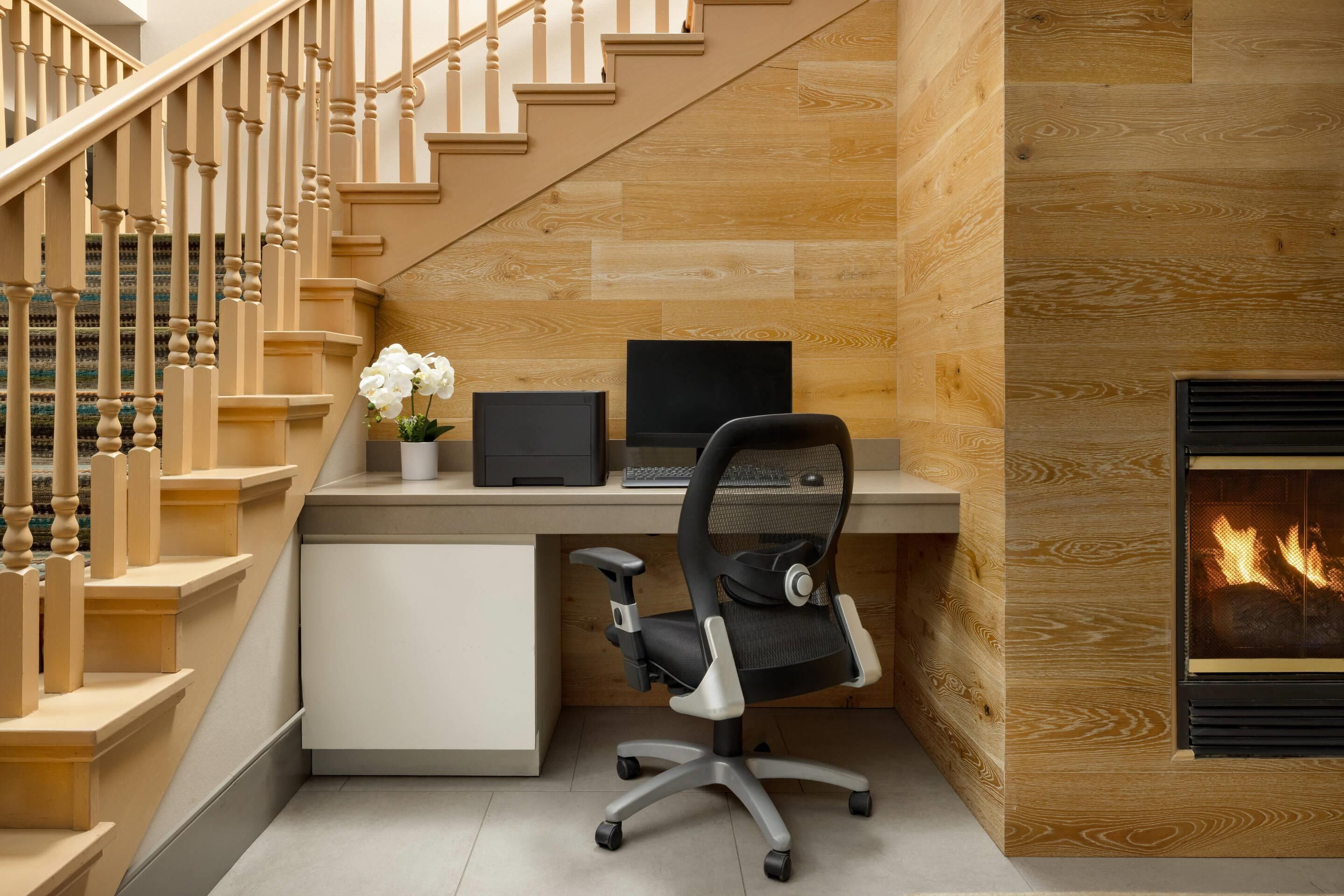 Ein modernes Homeoffice mit einer Holztreppe, einem Schreibtisch mit Laptop, einem schwarzen Bürostuhl und einem Kamin.