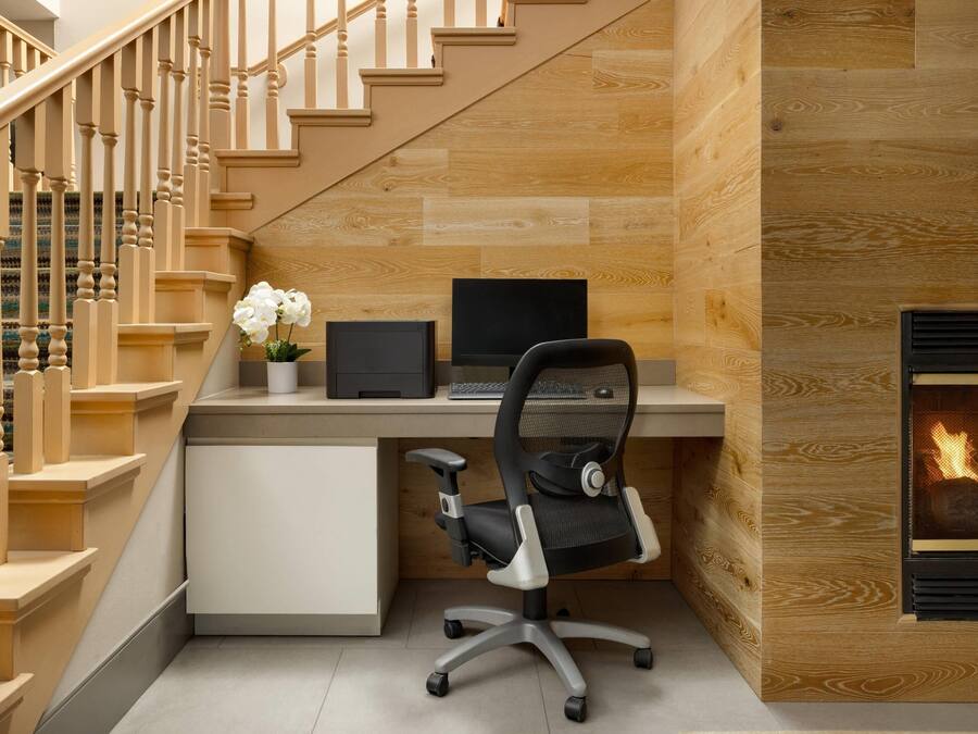 Una oficina moderna en casa con una escalera de madera, un escritorio con una computadora portátil, una silla de oficina negra y una chimenea.