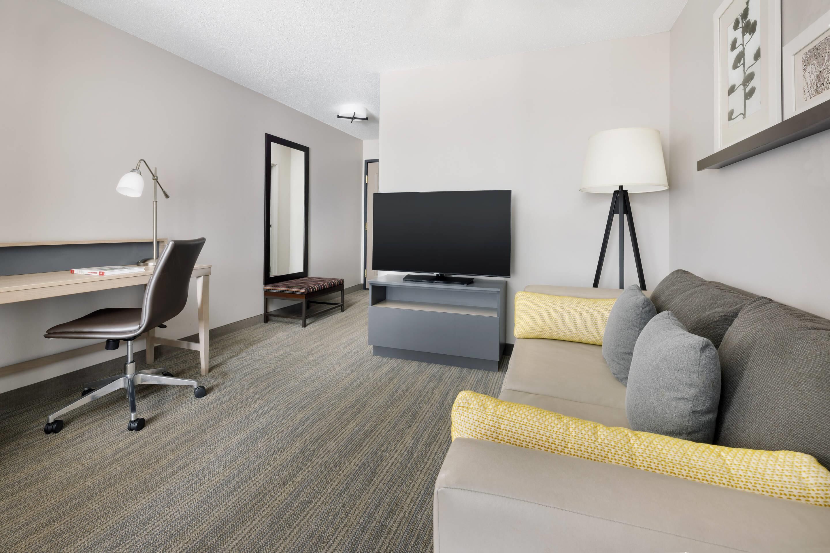 A modern hotel room with a desk, chair, television, sofa, and lamps.