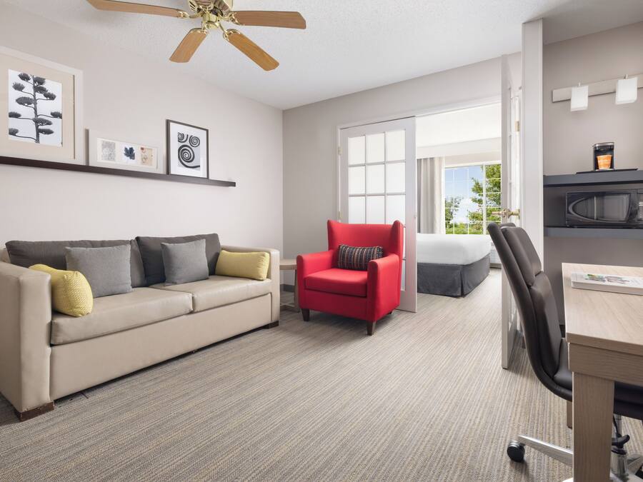 A modern hotel room with a sofa, a red armchair, a desk, a microwave, and a ceiling fan.