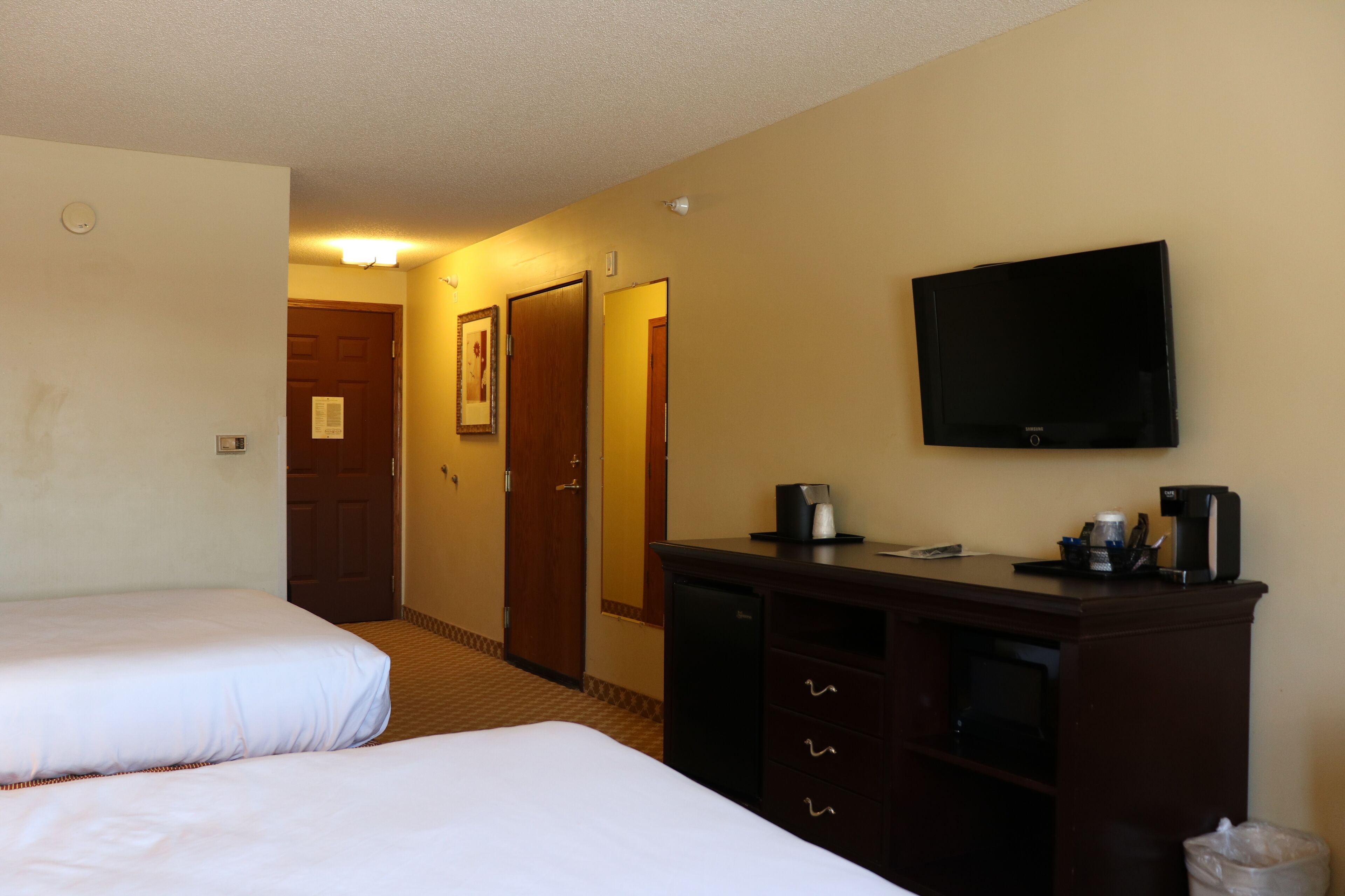 Habitación de hotel con dos camas, un escritorio con cafetera, televisor y una puerta que da a otra habitación.
