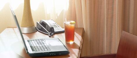 Habitación de hotel con una mesa de madera, una computadora portátil, un teléfono, un vaso de jugo y una lámpara.
