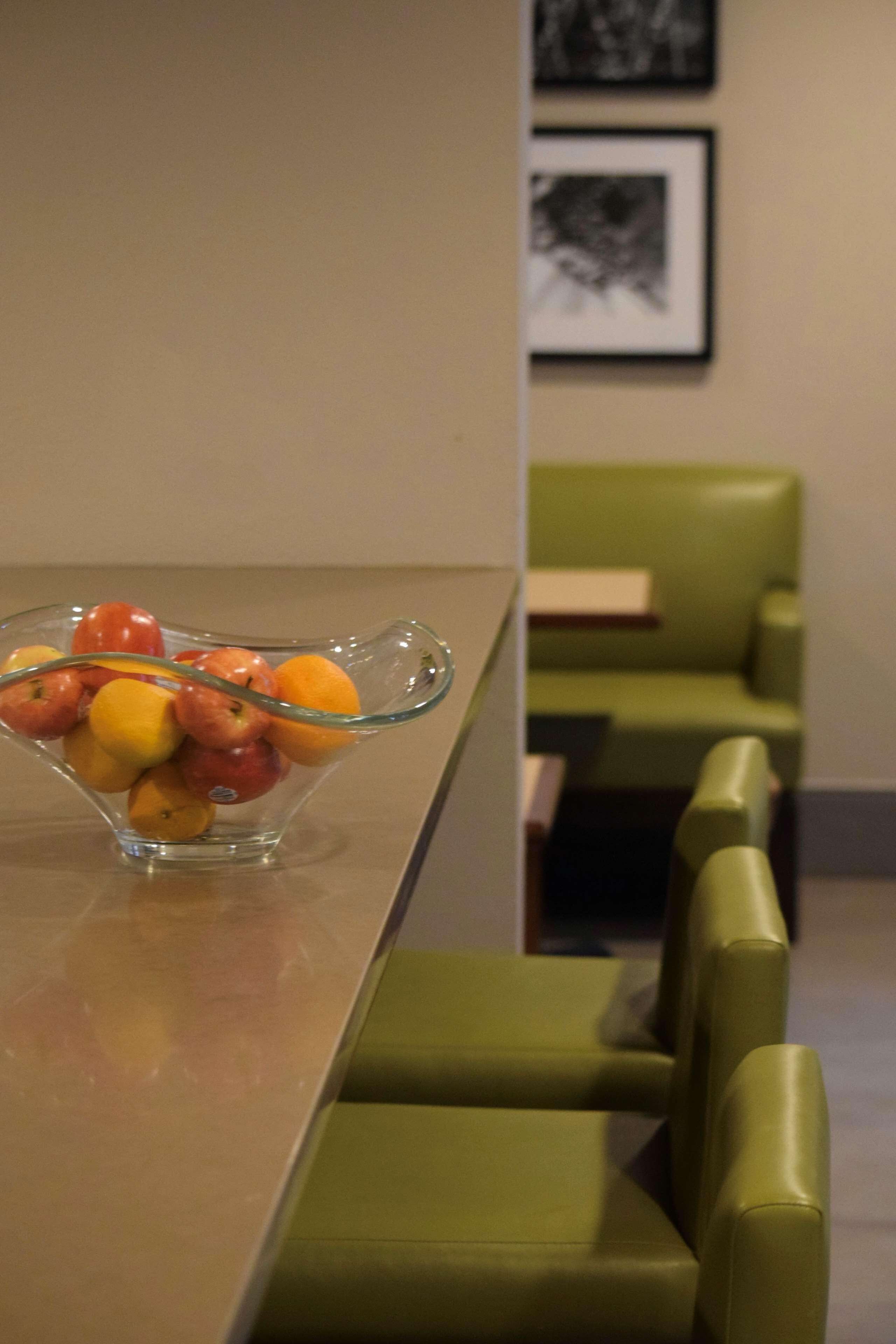 A clear glass bowl filled with fruits on a table, green upholstered chairs, and a framed picture on the wall.
